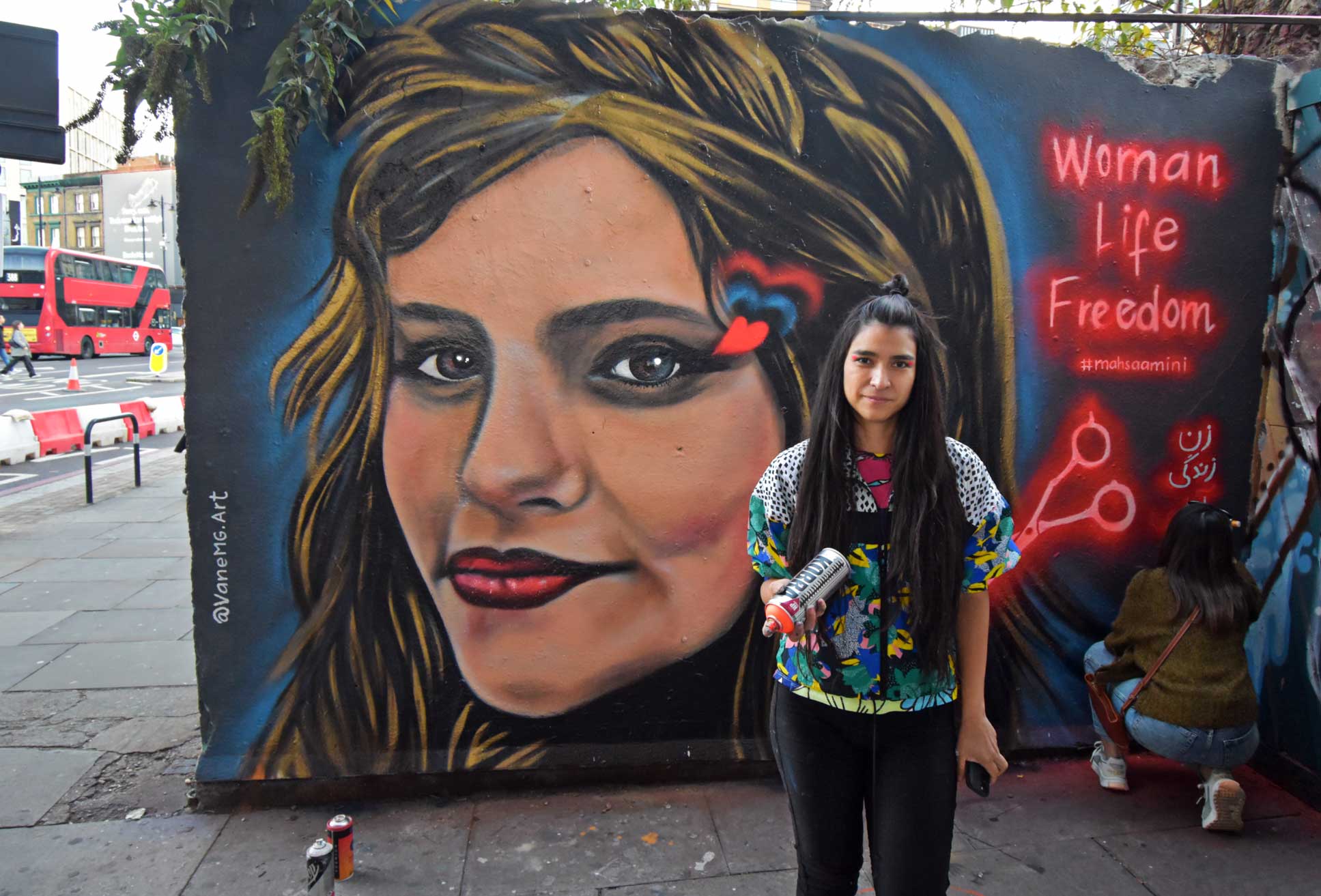 A young woman holding a spray paint bottle, stands proudly in front of her mural depicting the face of Mahsa Amini.