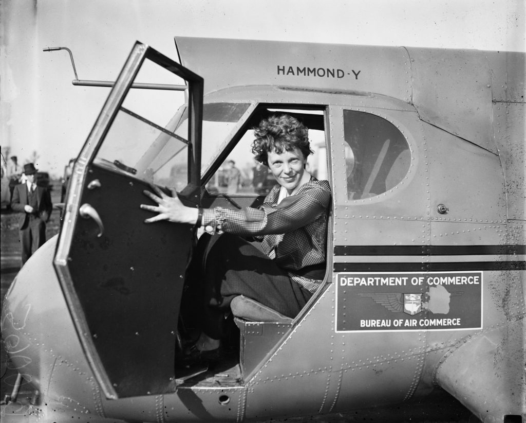 Vintage photo of Amelia Earheart sitting in the cockpit of an aircraft, smile at the camera through an open door.